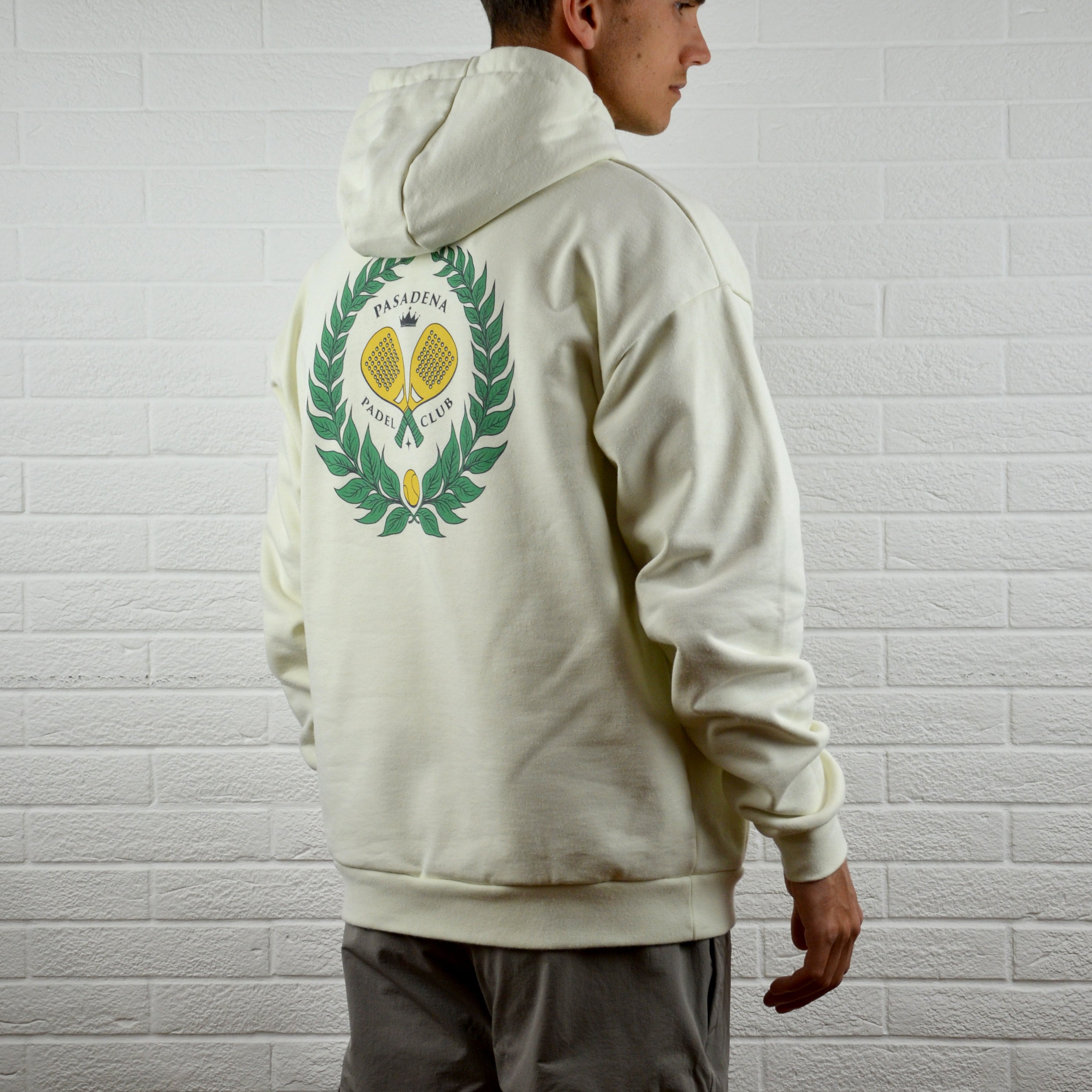 Person wearing a cream hoodie with a logo on the back against a white brick wall.